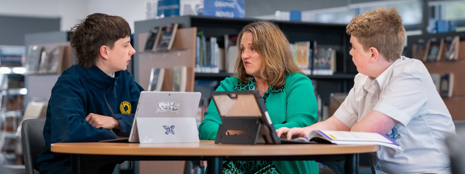 A teacher and two students sit with their laptops at a table in a library.